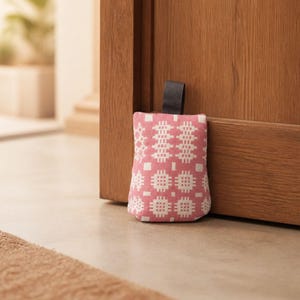 Puede incluir: Un tope de puerta de tela rectangular con un patr&oacute;n geom&eacute;trico rosa y blanco. El tope de puerta tiene una peque&ntilde;a asa de tela negra en la parte superior y est&aacute; colocado contra una puerta de madera para mantenerla fija en un suelo claro.