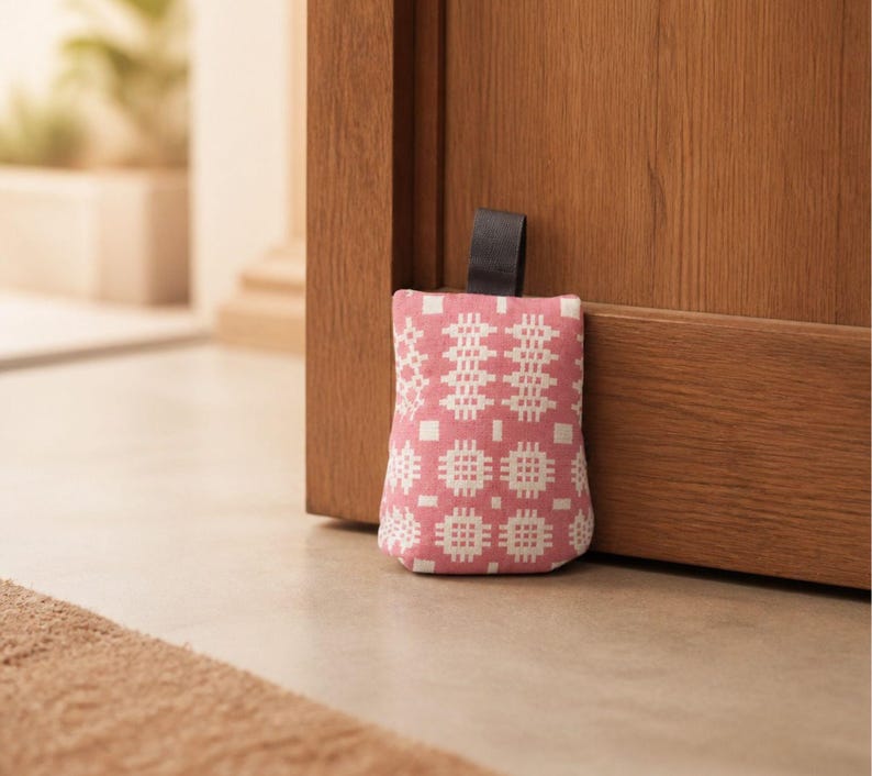Puede incluir: Un tope de puerta de tela rectangular con un patr&oacute;n geom&eacute;trico rosa y blanco. El tope de puerta tiene una peque&ntilde;a asa de tela negra en la parte superior y est&aacute; colocado contra una puerta de madera para mantenerla fija en un suelo claro.