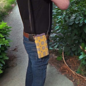 May include: A person wearing blue jeans and a black shirt with a yellow and gray patterned phone case attached to a black strap.