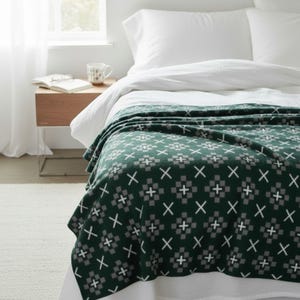 Puede incluir: Una manta de color verde oscuro con un patrón geométrico repetitivo de cuadrados grises y cruces blancas. La manta está colocada sobre una cama blanca con almohadas blancas, junto a una mesita de noche de madera con un libro y una taza.