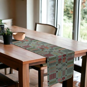 May include: A green table runner with a geometric pattern in shades of red, orange, yellow, and brown. The runner is on a wooden table with a bowl and a plant on the surface.
