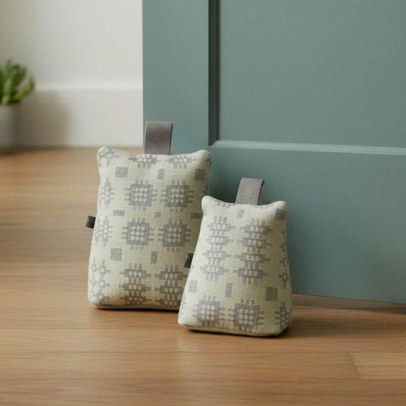 Puede incluir: Dos topes de puerta con una funda de tela beige claro que presenta un patr&oacute;n geom&eacute;trico gris repetitivo. Cada tope tiene una forma rectangular con un asa de tela gris unida al borde superior, dise&ntilde;ada para mantener la puerta abierta.