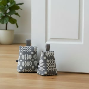 Puede incluir: Dos topes de puerta de tela gris con un patr&oacute;n geom&eacute;trico blanco. Cada tope tiene una forma rectangular, un asa superior y una peque&ntilde;a etiqueta de tela negra en el lateral. El patr&oacute;n consiste en cuadrados y dise&ntilde;os de rejilla repetidos.