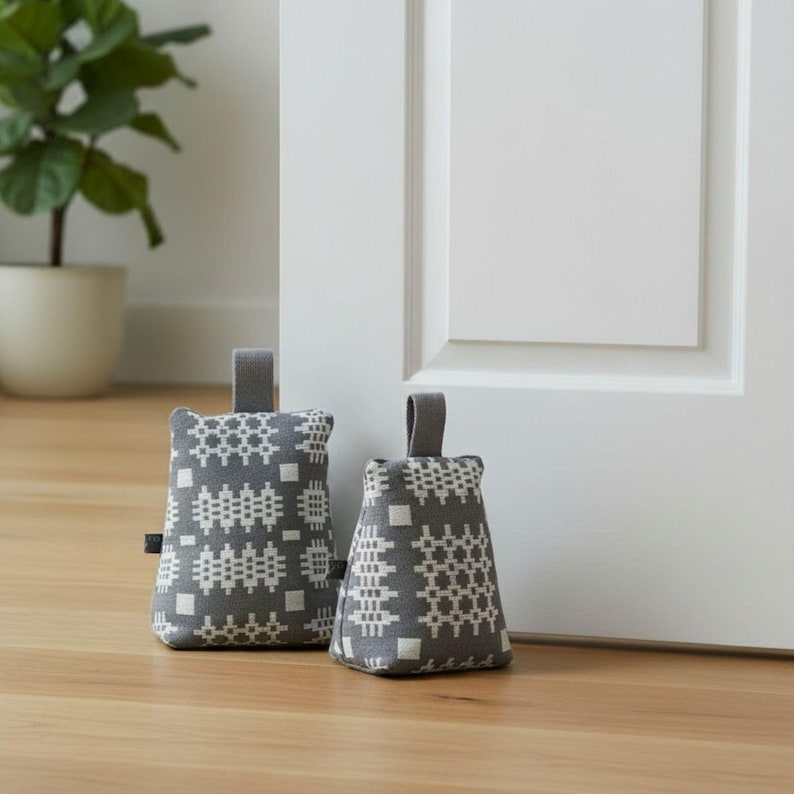Puede incluir: Dos topes de puerta de tela gris con un patr&oacute;n geom&eacute;trico blanco. Cada tope tiene una forma rectangular, un asa superior y una peque&ntilde;a etiqueta de tela negra en el lateral. El patr&oacute;n consiste en cuadrados y dise&ntilde;os de rejilla repetidos.