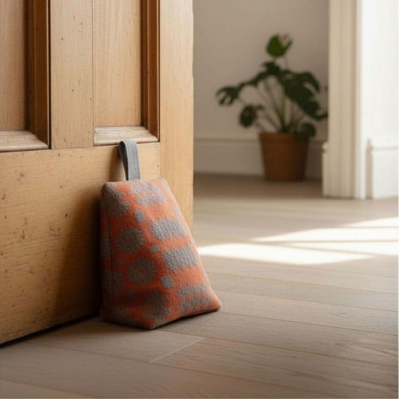 Puede incluir: Un tope de puerta en forma de cu&ntilde;a con un exterior de tela naranja vibrante y un patr&oacute;n texturizado gris repetitivo. El tope incluye un asa de tela gris resistente en la parte superior, dise&ntilde;ada para mantener una puerta de madera abierta.