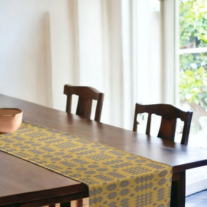 May include: A dining table with a mustard yellow and grey patterned table runner. Two dark wooden chairs are behind the table. A copper-coloured bowl sits on the table. A window with natural light is in the background.