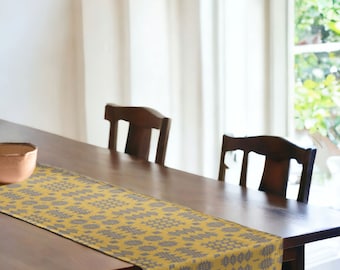 May include: A wooden dining table with a mustard yellow and gray patterned table runner. Two dark wooden chairs are positioned behind the table. A copper-colored bowl sits on the table. A window with natural light is in the background.