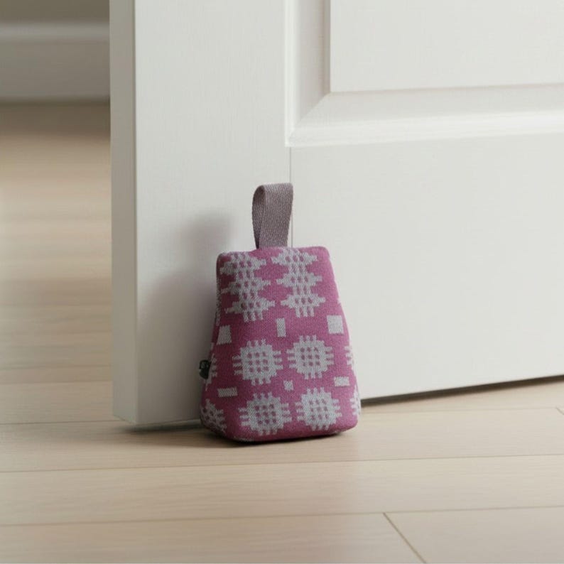 Puede incluir: Un tope de puerta en forma de cu&ntilde;a con un exterior de tela color magenta intenso y un patr&oacute;n geom&eacute;trico gris claro repetitivo. El tope cuenta con una peque&ntilde;a y resistente asa de tela en la parte superior central para facilitar su uso.