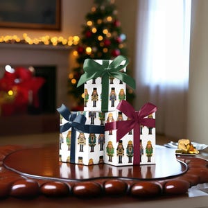 Könnte beinhalten: Drei verpackte Geschenkboxen mit weißem Hintergrund und einem Muster aus Spielzeug-Nussknackern. Jede Box ist mit einer unterschiedlich farbigen Schleife gebunden: Grün, Marineblau und Burgunderrot. Die Geschenke stehen auf einem dunkelbraunen Tisch, im Hintergrund ein Weihnachtsbaum.