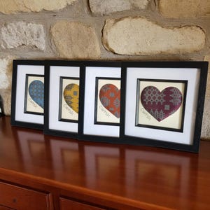 May include: Four framed heart-shaped artworks. Each heart is a different color: blue, yellow, orange, and burgundy, with a patterned design. The word "Cwtsh" is printed below each heart. The frames are black, and the background is a stone wall.