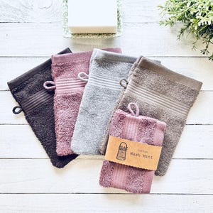 May include: Five different coloured washcloths, including brown, pink, grey, and a darker pink. The washcloths are stacked on top of each other and have a tag that says "Cotton Wash Mitt".