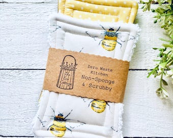 May include: Two white and yellow kitchen scrubbies with a bee pattern. The scrubbies are made of cotton and have a frayed edge. The scrubbies are packaged with a brown paper label that reads "Zero Waste Kitchen Non-Sponge & Scrubby".