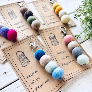 May include: Several felted wool keyrings in shades of pink, blue, and green. Each keyring has a series of wool balls attached to a silver-tone clasp. The keyrings are presented on kraft paper cards with the words "Felted Wool Keyring".