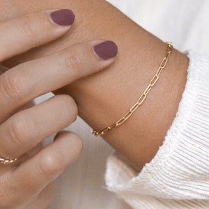 Könnte beinhalten: Ein goldenes Kettenarmband mit einem zarten Gliederdesign. Das Armband wird am Handgelenk einer Person getragen, mit zwei goldenen Ringen an derselben Hand.