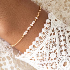 Könnte beinhalten: Ein zartes goldenes Armband mit kleinen, weißen Perlen. Das Armband wird an einem Handgelenk getragen, mit einem detaillierten weißen Spitzensaum im Hintergrund. Der Schmuck ist ein schlichtes, aber elegantes Accessoire.