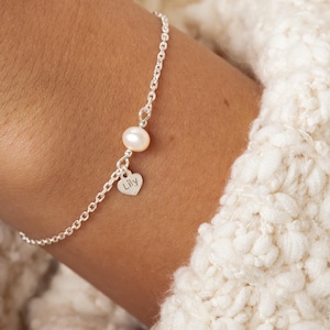 Könnte beinhalten: Zartes Silberarmband mit einer kleinen Perle und einem herzförmigen Anhänger mit dem Namen "Lily". Das Armband wird an einem Handgelenk vor einem weichen, strukturierten cremefarbenen Hintergrund gezeigt.