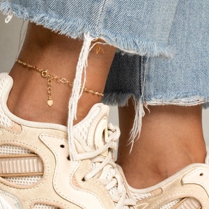 Könnte beinhalten: Ein goldenes Fußkettchen mit einem kleinen Herz-Anhänger. Das Fußkettchen wird an einem Fuß mit weißen Turnschuhen und einer blauen Jeans getragen.