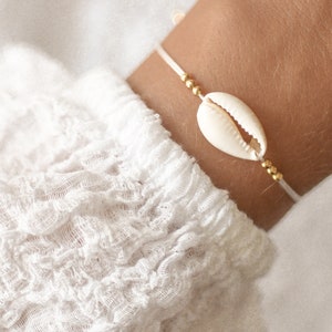 Könnte beinhalten: Ein Armband mit einem weißen Muschel-Anhänger und goldenen Perlen auf einer weißen Schnur.