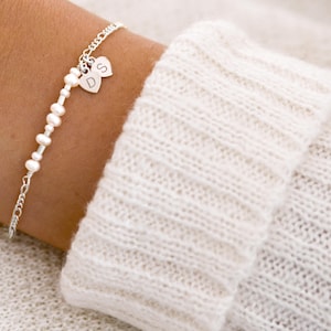 Könnte beinhalten: Ein filigranes Silberarmband mit Perlen- und Perlendesign. Zwei kleine Herzanhänger mit den Buchstaben "D" und "S" hängen an dem Armband. Das Armband wird an einem Handgelenk getragen, neben einem weißen Strickärmel.