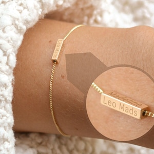 Puede incluir: Una pulsera dorada con una cadena fina. La pulsera presenta un colgante rectangular grabado con los nombres "Leo Mads" y "Madeline". La pulsera se lleva en la muñeca.