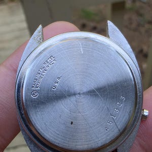 May include: Close-up of the back of a vintage wristwatch. The watch has a stainless steel back with the text "BASE METAL BEZEL STAINLESS BACK U.S.A." and the number "451323". The watch band is silver.