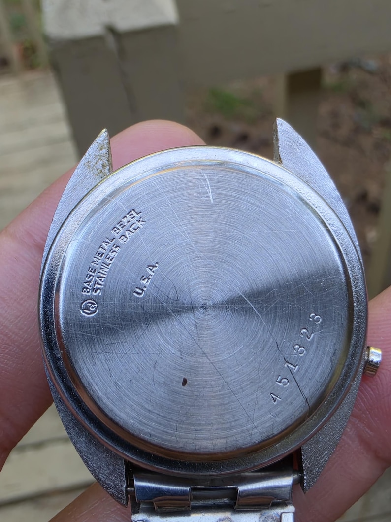 May include: Close-up of the back of a vintage wristwatch. The watch has a stainless steel back with the text "BASE METAL BEZEL STAINLESS BACK U.S.A." and the number "451323". The watch band is silver.