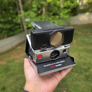 Peut inclure: Un appareil photo Polaroid SX-70 Land Camera vintage, tenu dans une main. L'appareil photo est argenté et noir avec un bouton rouge et un haut-parleur doré. Le texte "POLAROID SX-70 LAND CAMERA SONAR OneStep" est imprimé.