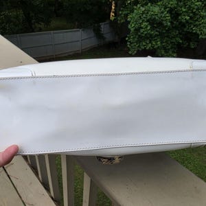 Op de afbeelding: Een witte rechthoekige handtasbodem van glad wit materiaal met zichtbare stiksels langs de randen. Het onderste paneel is plat en langwerpig en toont een cirkelvormig metalen hardware-detail dat in het midden van de onderrand is geplaatst.