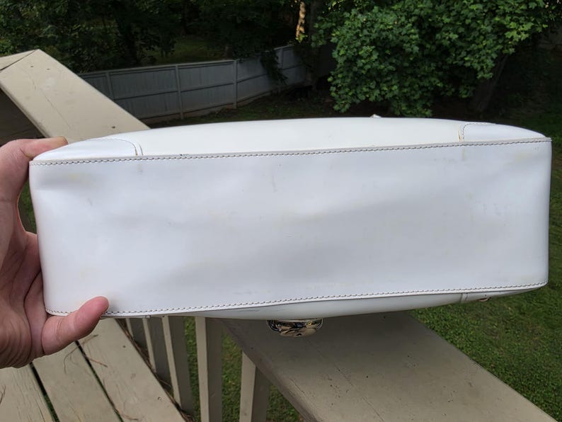Op de afbeelding: Een witte rechthoekige handtasbodem van glad wit materiaal met zichtbare stiksels langs de randen. Het onderste paneel is plat en langwerpig en toont een cirkelvormig metalen hardware-detail dat in het midden van de onderrand is geplaatst.