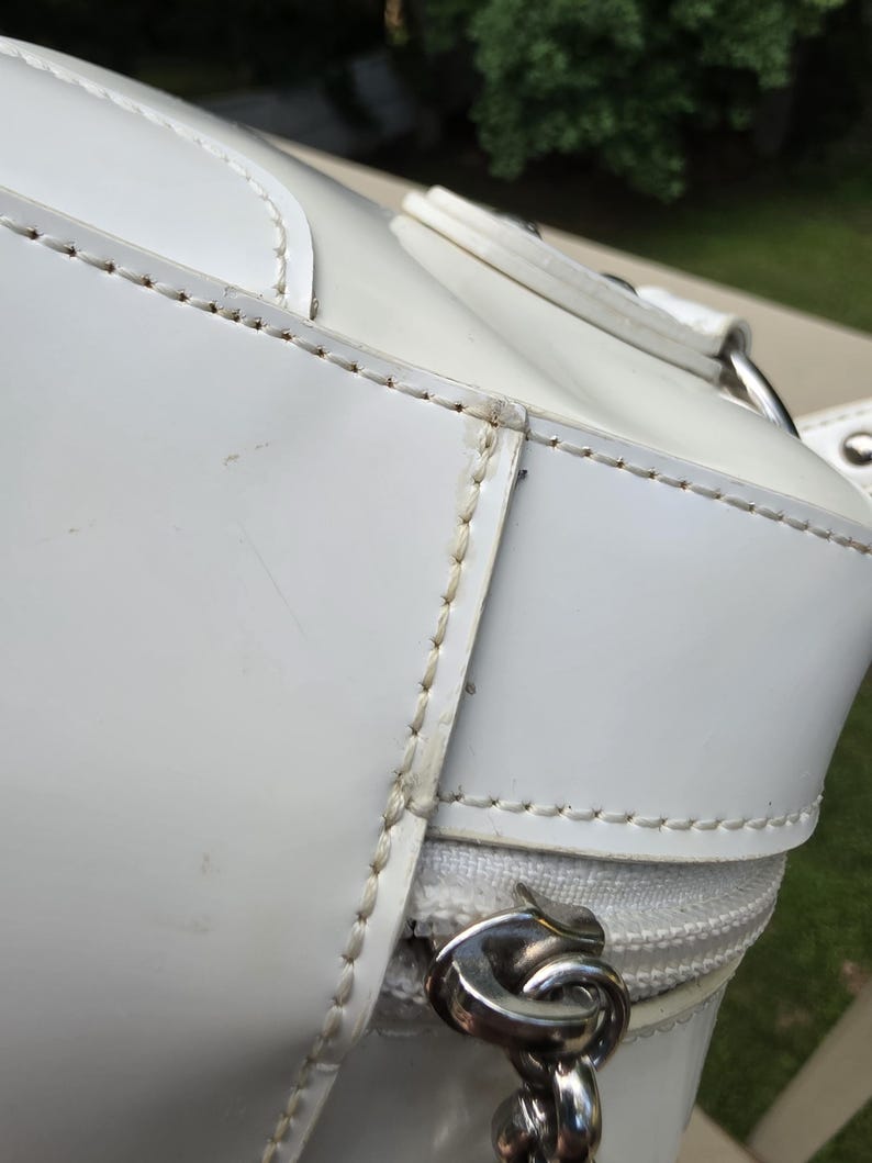 Op de afbeelding: Een witte handtas met gladde panelen en zichtbare witte stiksels langs de naden. De tas is voorzien van een witte ritssluiting met een zilverkleurige metalen trekker en een zilverkleurige ketting bevestigd aan het zijbeslag.