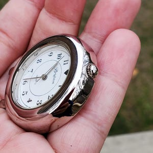 May include: A silver-toned Brighton quartz pocket watch with a white face and black numerals. The watch has an oval shape and a small crown for winding. The watch is held in a hand.