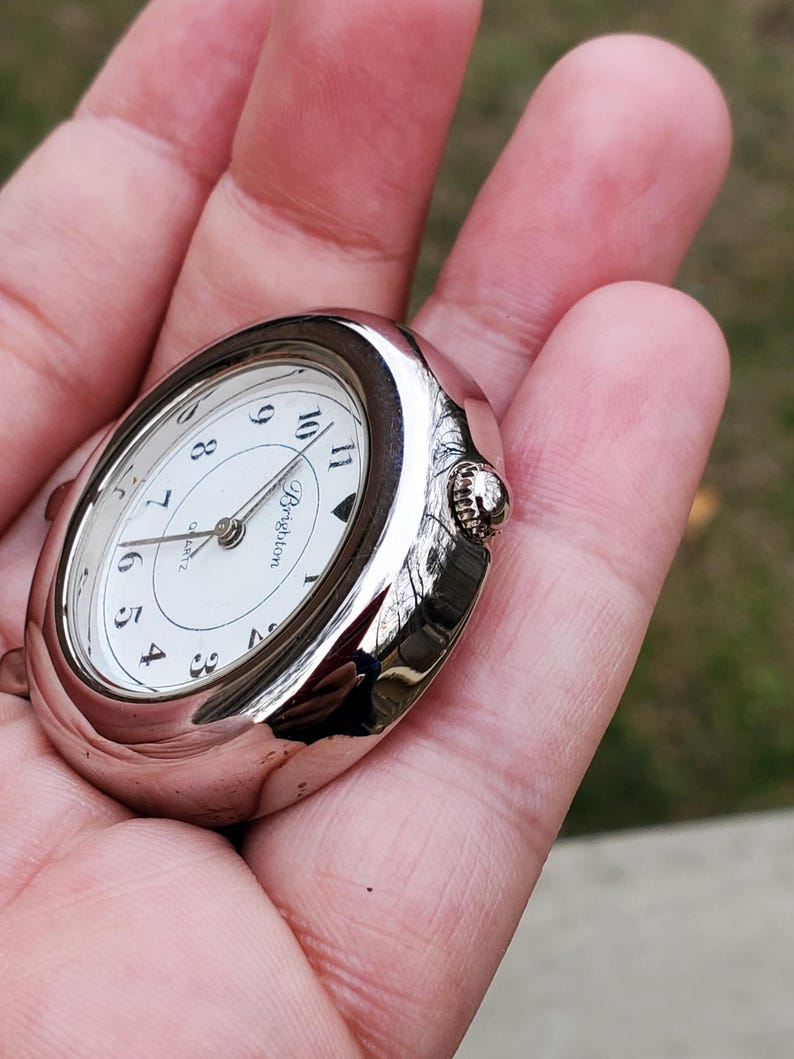 May include: A silver-toned Brighton quartz pocket watch with a white face and black numerals. The watch has an oval shape and a small crown for winding. The watch is held in a hand.
