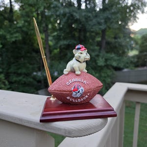 May include: A desk pen holder featuring a brown football with the text GEORGIA BULLDOGS and a team logo. A white bulldog figurine wearing a red and black cap sits on top of the football. A gold-toned pen is inserted into a holder on the side of the base.