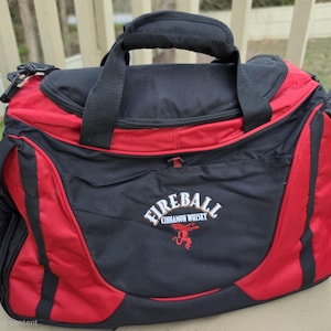 K&ouml;nnte beinhalten: Eine schwarz-rote Reisetasche mit oberem Griff und Schultergurt. Auf der Tasche steht in Wei&szlig; der Schriftzug "Fireball Cinnamon Whisky" mit einem roten Logo. Die Tasche liegt auf einer Holzoberfl&auml;che.