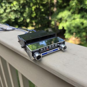 May include: A vintage Audiovox FM radio receiver featuring a black metal chassis, a chrome-finished front panel with a tuning scale ranging from 88 to 108 MHz, two circular control knobs, and blue accent detailing with exposed electrical wiring.