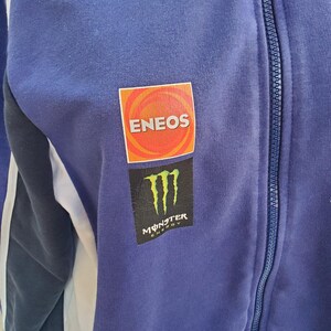 May include: A blue zip-up jacket with white stripes on the sleeves. The jacket features the ENEOS logo in a red and orange square and the Monster Energy logo in black and green.
