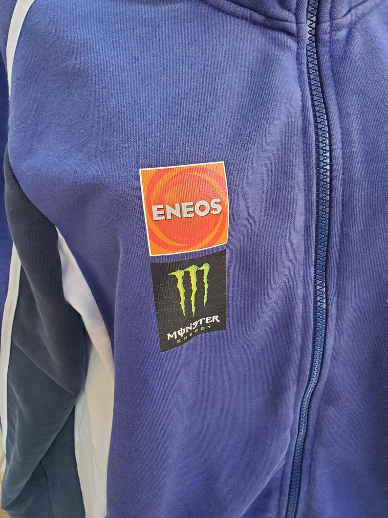 May include: A blue zip-up jacket with white stripes on the sleeves. The jacket features the ENEOS logo in a red and orange square and the Monster Energy logo in black and green.