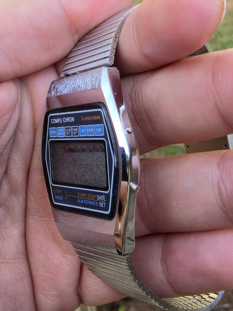 Może przedstawiać: Zegarek cyfrowy Compuchron w stylu vintage ze srebrnym metalowym paskiem. Tarcza zegarka jest czarna z cyfrowym wyświetlaczem i przyciskami funkcyjnymi. Widoczny jest napis "COMPU CHRON" i "ALARMCHRONO".
