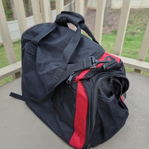 K&ouml;nnte beinhalten: Schwarz-rote Reisetasche mit Schultergurt und Griff. Die Tasche hat ein Hauptfach mit Rei&szlig;verschluss und eine Seitentasche mit Mesh-Einsatz. Die Tasche ist aus strapazierf&auml;higem Material gefertigt und f&uuml;r Reisen oder Sport konzipiert.
