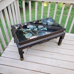 May include: Black rectangular coffee table with a glass top featuring a floral design in shades of white, green, and blue. The table has dark brown legs with gold accents and is placed on a wooden deck.