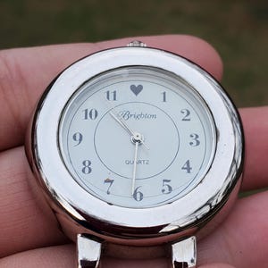 May include: A silver-toned Brighton quartz watch face with a white dial. The watch face has black numerals, a heart symbol above the 12, and the brand name "Brighton" in script. The watch has two small feet.