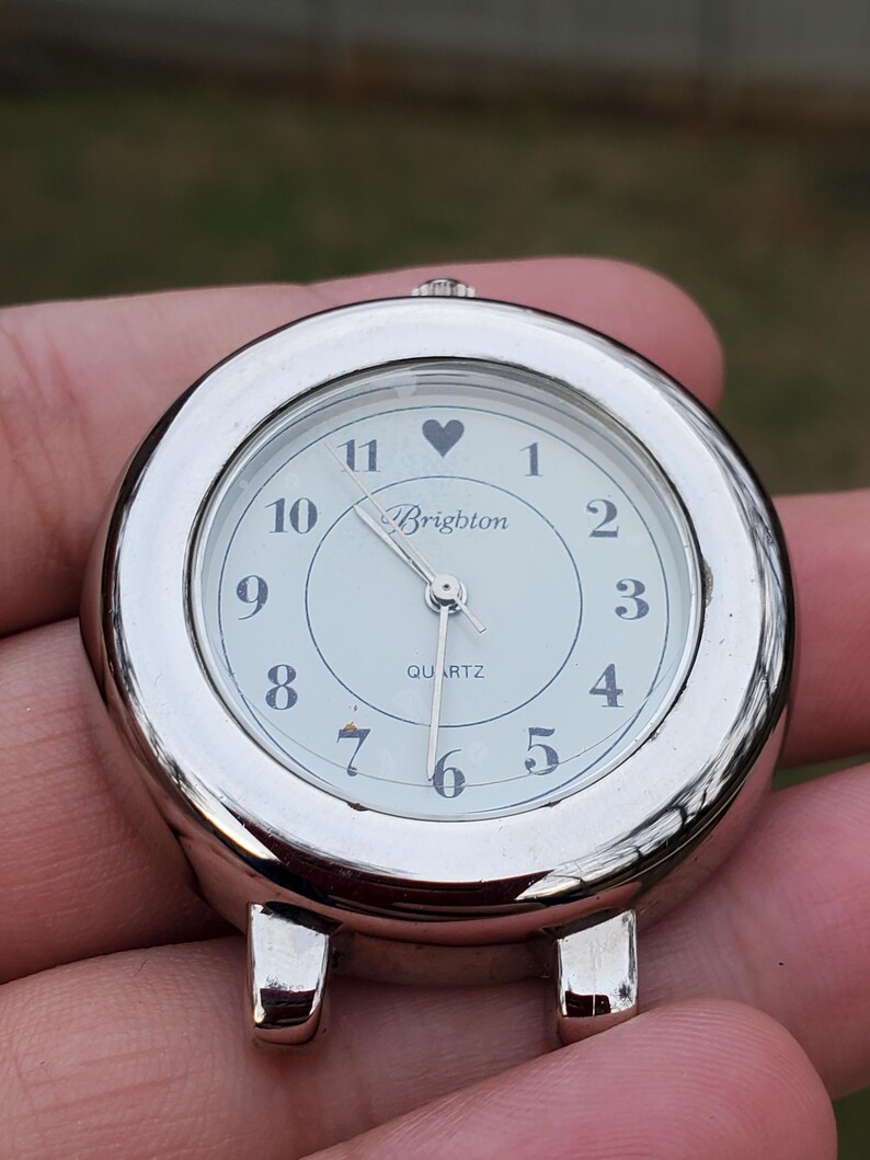 May include: A silver-toned Brighton quartz watch face with a white dial. The watch face has black numerals, a heart symbol above the 12, and the brand name "Brighton" in script. The watch has two small feet.