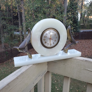 May include: A vintage clock with a white face and gold-colored hands and numerals. The clock is set in a white, circular frame and rests on a white rectangular base. Bronze-colored bird sculptures flank the clock, adding a decorative touch.
