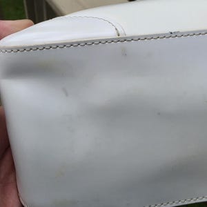 Op de afbeelding: Een witte handtas met een gladde, glanzende afwerking en zichtbare stiksels langs de bovennaad. Het oppervlak vertoont lichte gebruikssporen, waaronder een lichte verkleuring en kleine krasjes, met een gestructureerde vorm.