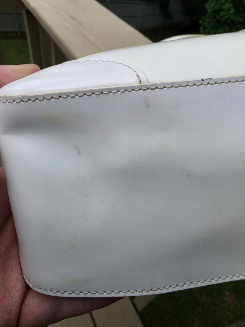 Op de afbeelding: Een witte handtas met een gladde, glanzende afwerking en zichtbare stiksels langs de bovennaad. Het oppervlak vertoont lichte gebruikssporen, waaronder een lichte verkleuring en kleine krasjes, met een gestructureerde vorm.