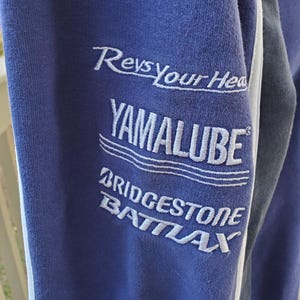 May include: Close-up of a blue sweatshirt sleeve with white embroidered text. The text includes "Revs Your Head," "YAMALUBE," "BRIDGESTONE," and "BATLAX." A white stripe runs along the edge of the sleeve.