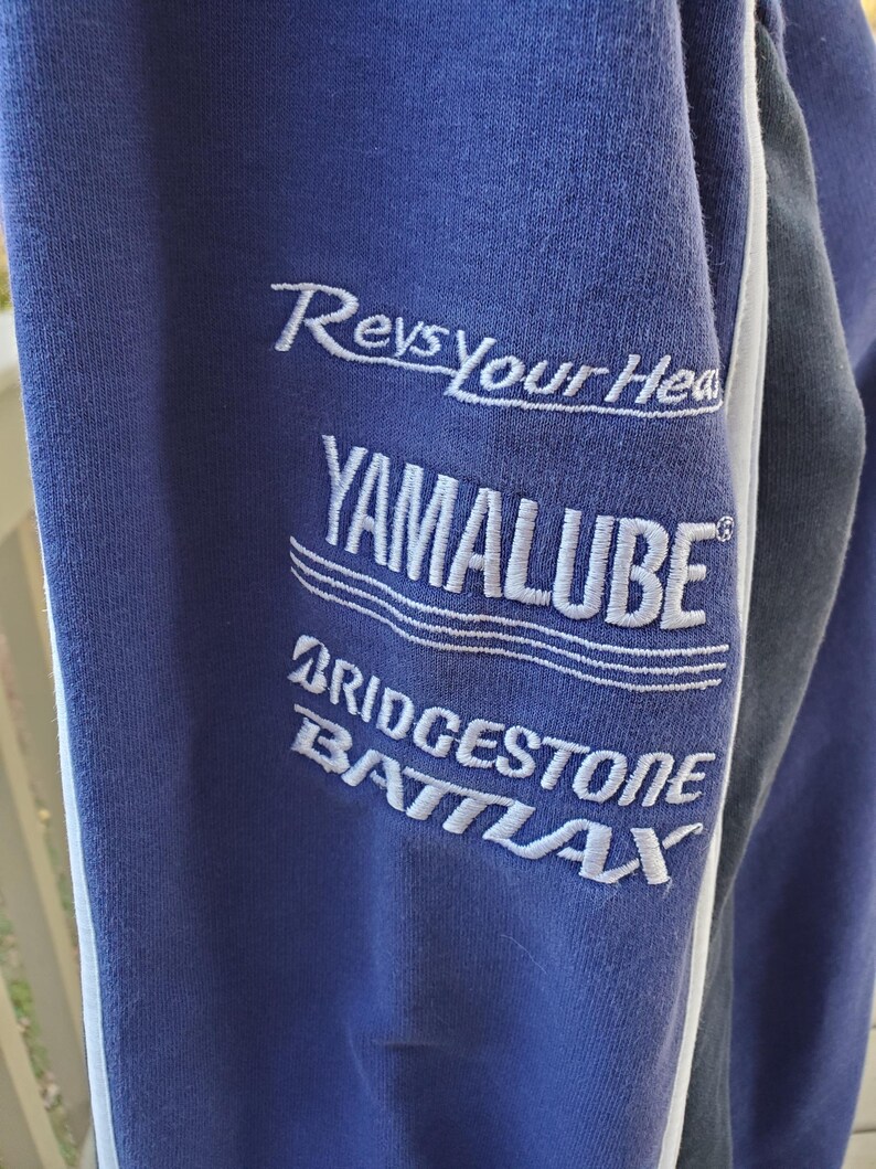 May include: Close-up of a blue sweatshirt sleeve with white embroidered text. The text includes "Revs Your Head," "YAMALUBE," "BRIDGESTONE," and "BATLAX." A white stripe runs along the edge of the sleeve.