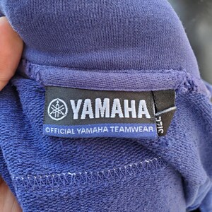 May include: Close-up of a blue sweatshirt with a tag that reads "YAMAHA OFFICIAL YAMAHA TEAMWEAR". The tag features the Yamaha logo, a stylized emblem. The sweatshirt's fabric has a textured appearance.