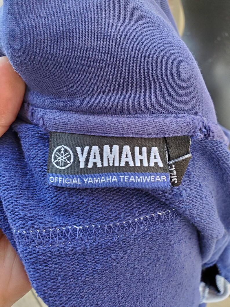 May include: Close-up of a blue sweatshirt with a tag that reads "YAMAHA OFFICIAL YAMAHA TEAMWEAR". The tag features the Yamaha logo, a stylized emblem. The sweatshirt's fabric has a textured appearance.