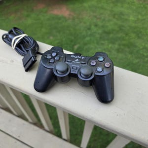 May include: A black Sony PlayStation wired game controller featuring two analog sticks, a directional pad, four action buttons with geometric symbols, and central Select, Start, and Analog buttons. The controller includes a black cable with a connector.